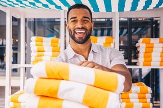 A joyful man stands in front of neatly stacked orange and white towels, exuding warmth and friendliness in a bright, sunny setting.