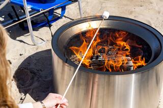 A person roasts a marshmallow over a blazing fire in a stylish metal fire pit, with a blue camping chair nearby on the sandy ground.