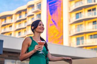A woman in a green dress smiles while holding a cocktail, set against a vibrant building backdrop during sunset.