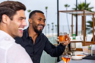 Two men enjoy drinks and share a laugh at a stylish venue with palm trees in the background, capturing a moment of friendship and joy.