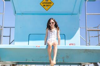 A girl in sunglasses sits on a lifeguard stand by the beach, smiling under a clear sky with a "No Lifeguard On Duty" sign above her.