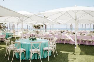 A beautifully arranged outdoor event space features round tables with mint and pink linens, surrounded by umbrellas and ocean views.