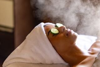 A person relaxes with a towel wrapped around their head and cucumber slices over their eyes, surrounded by a soothing mist.