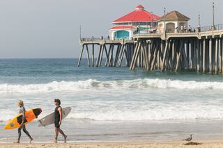 Two surfers stroll along a sandy beach with surfboards, while a colorful pier and gentle waves create a relaxing coastal scene.