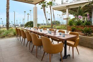 A long wooden dining table with stylish chairs is set up outdoors, surrounded by palm trees and a view of the sea.