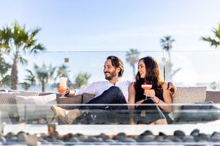 A joyful couple relaxes on a stylish patio, sipping colorful drinks against a backdrop of palm trees and sunny skies.