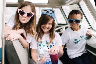 Three kids, stylishly dressed in matching t-shirts and vibrant sunglasses, enjoy a fun moment inside a retro vehicle, exuding joy and friendship.