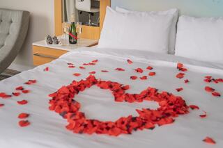 A romantic setup features a heart made of red rose petals on a bed, complemented by a bottle of champagne and glasses nearby.