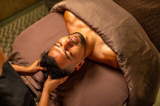 A man relaxes with his eyes closed while receiving a head massage, covered by a brown blanket in a spa-like setting.