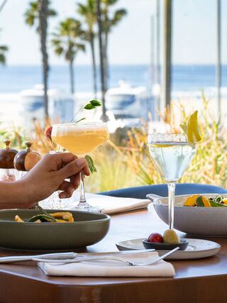 Two cocktails sit on a table with a scenic ocean view, palm trees in the background, and a vibrant meal waiting to be enjoyed.