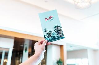 A hand holds a brochure featuring palm trees and the text "Pacific City," set against a bright, airy background.