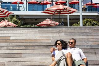 A couple enjoys a sunny day outdoors, sitting together on wooden benches with red and white parasols in the background.