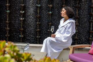A woman in a white robe sits peacefully by a decorative wall, holding a glass of champagne, enjoying a moment of relaxation.