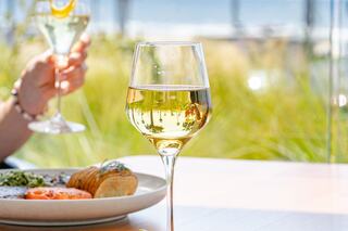 A glass of white wine rests on a table, reflecting greenery. In the background, a hand holds another drink, enhancing a relaxing dining scene.