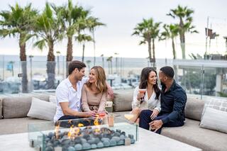 A lively gathering of four friends enjoying drinks by a cozy fire pit, surrounded by palm trees and a beach view. Relaxation and laughter abound.