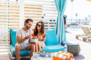 A couple enjoys a cozy moment together, sharing food and drinks by a poolside, surrounded by palm trees and bright sunshine.