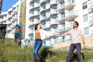 A couple joyfully holds hands while walking through grass, with a modern building and playful decor in the background.