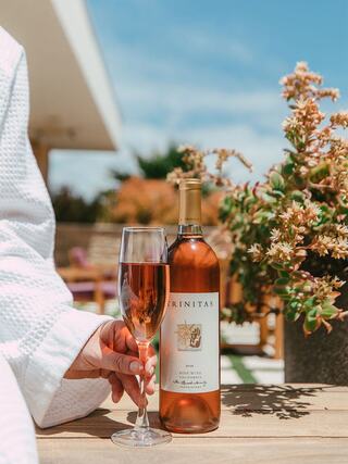 A glass of rosé wine is held beside a bottle labeled "Trinitas," with lush greenery and a sunny outdoor setting in the background.