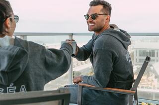 Two people in matching hoodies enjoy a sunny moment on a balcony, sharing a drink and smiles against a scenic backdrop.