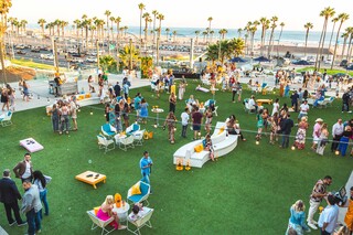 A lively outdoor gathering on green grass, with people socializing, enjoying drinks, and a backdrop of palm trees and the ocean.