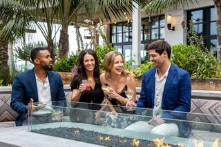 Four friends enjoy cocktails, laughing together by a fire pit surrounded by greenery in a stylish outdoor setting.