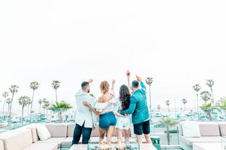 A group of four friends toasts together on a sunny terrace, overlooking palm trees and a beach, capturing a moment of celebration and friendship.