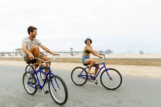 A man and woman joyfully ride blue bicycles along a sandy beach path, enjoying a fun day by the seaside under a cloudy sky.