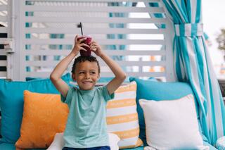 A cheerful boy with curly hair raises a drink above his head, sitting on a vibrant, cozy couch decorated with colorful pillows.