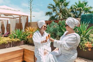 Two women in robes and towels enjoy drinks while relaxing in a lush, sunny spa-like setting with palm trees and vibrant greenery.