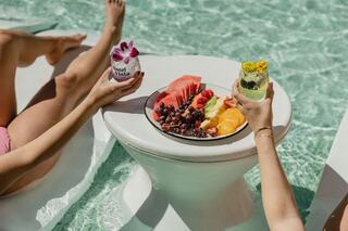 Two hands hold colorful drinks above a fruit platter on a table by the pool, inviting relaxation and enjoyment on a sunny day.
