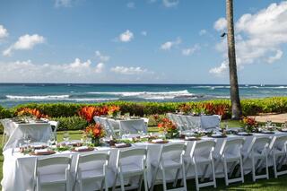 A beautifully set long dining table overlooking the ocean, surrounded by lush greenery and vibrant tropical flowers under a blue sky.