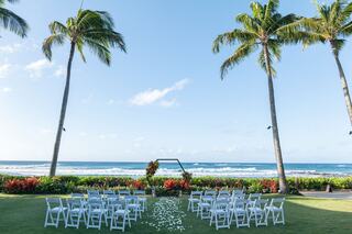 A picturesque outdoor wedding setup by the ocean, featuring white chairs, tropical palm trees, and colorful floral decorations.
