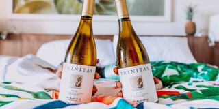 Two bottles of Trinitas Chardonnay are held up in bed, surrounded by colorful bedding and a relaxing ambiance.