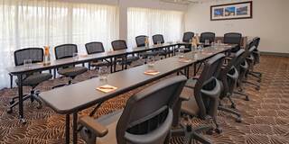 A modern conference room with long tables, black chairs, and natural light filtering through sheer curtains, designed for meetings or events.