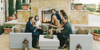 A group of six people enjoys drinks and conversation by a stone fireplace in an outdoor setting, surrounded by greenery and cozy furniture.