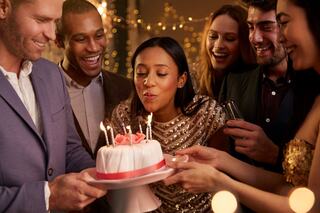 A woman joyfully blows out candles on a cake, surrounded by smiling friends celebrating a special occasion.