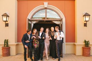 A stylish group of six friends exits a building, dressed elegantly and smiling, embodying joy and celebration in a vibrant outdoor setting.
