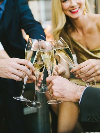 Glasses filled with sparkling beverage clink together in celebration, surrounded by elegantly dressed individuals sharing joyful moments.