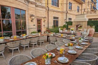 A beautiful outdoor dining setup features a long wooden table adorned with floral centerpieces, set against a stylish stone backdrop and a cozy fireplace.