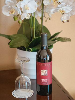A bottle of Trinitas wine sits beside an elegant glass, both placed on a polished surface with lush white orchids in the background.
