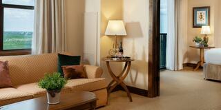 A cozy, well-lit hotel room features a sofa, coffee table, and lamps, with a view through the window and adjoining bedroom area.