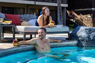 Two people relax by a pool, enjoying drinks. One is lounging on a chair, while the other sits in the water, smiling and soaking up the sun.