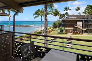 A tropical scene featuring palm trees, a sandy beach, and ocean waves, with a balcony view overlooking lush greenery and lounge chairs.