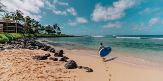A person walks along a sandy beach, carrying a blue surfboard, with palm trees and clear water in the background under a bright sky.