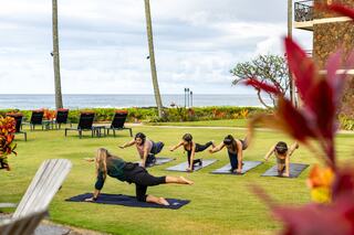A group of people practices yoga outdoors on mats, surrounded by lush greenery and stunning ocean views under a cloudy sky.