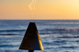 A metal cone emits a gentle wispy smoke against a serene ocean backdrop, illuminated by the warm hues of a sunset.