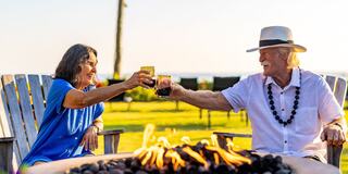 A joyful moment shared between an older man and woman, toasting with drinks by a fire pit, against a scenic outdoor backdrop.