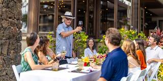 A man raises a glass in toast at a festive outdoor gathering, surrounded by smiling friends enjoying a colorful meal and drinks.