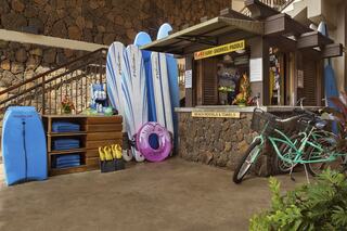 A beach rental kiosk features surfboards, boogie boards, and towels, alongside a bicycle, set against a stone wall and tropical decor.
