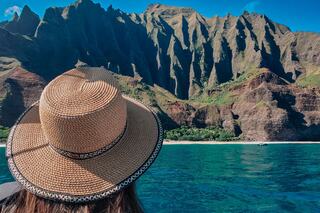 A person wearing a large straw hat gazes at dramatic green cliffs rising above sparkling blue waters, creating a serene coastal scene.
